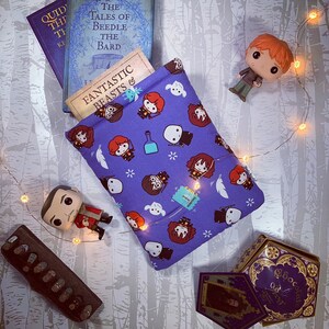 Può includere: Una custodia per libri viola con un motivo ripetuto di personaggi dei cartoni animati e icone, che contiene tre libri. I libri sono intitolati "Fantastic Beasts &", "The Tales of Beedle the Bard" e "Quidditch Through the Ages".