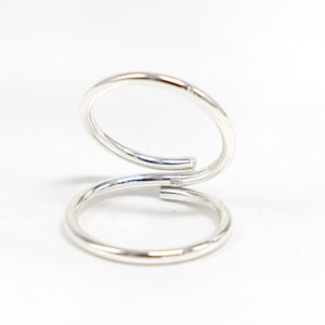 May include: A silver-colored ring with a unique double-band design. The ring features two circular bands that are connected, creating a modern and minimalist aesthetic. The bands are smooth and shiny, reflecting light.