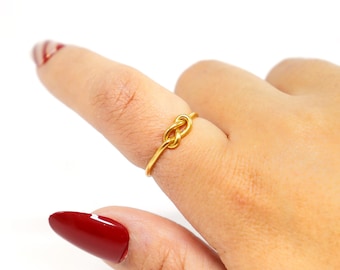 May include: A gold-coloured ring in a knot design is worn on a finger. The ring is simple and elegant, with a polished finish. The background is white, highlighting the ring's design. The ring is a piece of jewellery.