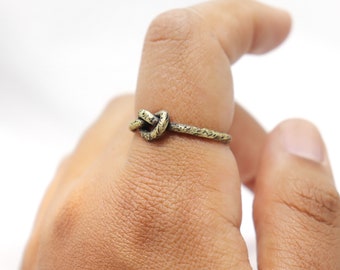 Puede incluir: Un anillo de color bronce con un diseño de nudo. El anillo se lleva en un dedo, mostrando los intrincados detalles del nudo y la banda texturizada. El metal del anillo tiene una apariencia ligeramente envejecida.
