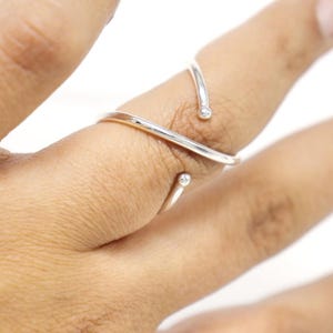 May include: A silver-colored ring with a unique, open design. The ring features two small, clear gemstones and wraps around two fingers. The ring is shown on a hand against a white background.