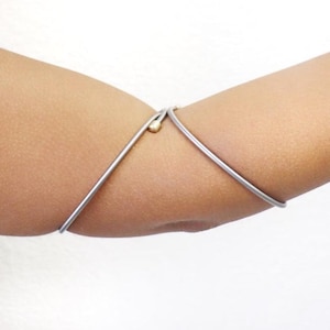 May include: A silver-toned metal arm cuff with a small gold-colored bead accent. The cuff is a simple, minimalist design, worn on the arm. The jewelry is shown against a neutral background.