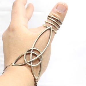 May include: A bronze-colored hand chain with a Celtic knot design. The chain features a series of rings for the thumb and a decorative element with a central knot and elongated teardrop shapes. A delicate chain connects the design to the wrist.