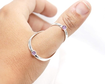 May include: A silver ring with two bands, each adorned with a small, round, purple gemstone. The ring is designed to be worn across two fingers. The gemstones are set in a simple, elegant style.