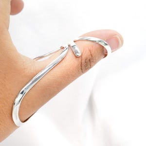 May include: A silver-colored ring designed to be worn on a thumb. The ring has a unique, open design with curved lines that wrap around the finger. The ring is made of a shiny, reflective metal.