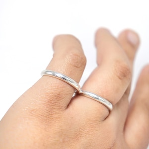 May include: Two silver rings are worn on fingers, set against a white background. The rings are simple bands, reflecting light. The image focuses on the jewelry, highlighting the design and material.
