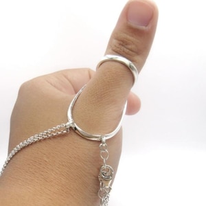 May include: A silver hand chain featuring a ring and a connecting chain. The ring is worn on the thumb, and the chain drapes down, connecting to a clasp. The jewelry is made of a shiny, metallic material.