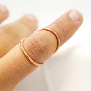 May include: Two thin, gold-colored rings are worn on a finger. The rings are simple bands, likely made of metal, and are positioned close together. The finger is against a white background, highlighting the jewelry.