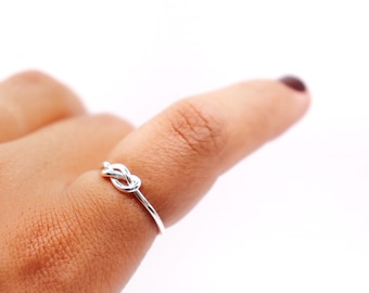 Könnte beinhalten: Ein zarter Silberring mit einem Knoten-Design. Der Ring ist dünn und sitzt auf einem Finger, der das komplizierte Knotendetail zeigt. Die polierte Oberfläche des Rings reflektiert das Licht.