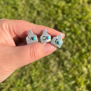 May include: Three small, silver-colored metal tags with turquoise-colored accents. Each tag has a different letter: M, C, and T. The tags are held in a hand against a blurred green background.