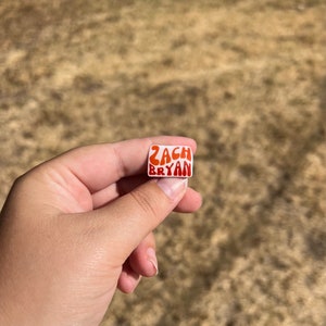 May include: A small, rectangular pin with the text "ZACH BRYAN" in orange, retro-style lettering. The pin is held between fingers against a blurred, brown background.