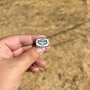 May include: A white enamel pin with a black border and a black and white bull's head design. The pin says "Wallen" in black letters.