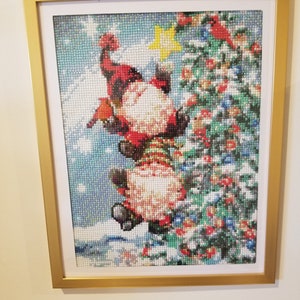 May include: A framed diamond painting depicting a gnome climbing a Christmas tree. The gnome is wearing a red hat and a white beard. The tree is adorned with red berries and a yellow star.