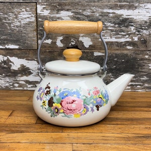Puede incluir: Tetera de esmalte blanco con un diseño floral con flores rosas, azules, amarillas y moradas. La tetera tiene un asa de madera y una tapa plateada.