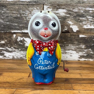 May include: A vintage wind-up toy of Peter Cottontail the rabbit. The toy is made of metal and has a gray and white rabbit head with a red and white polka dot bow tie. The rabbit is wearing blue overalls with yellow accents and the words "Peter Cottontail" are printed on the front. The toy has a red wind-up key on the side.