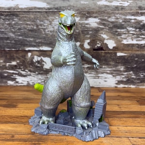May include: A silver Godzilla figurine with yellow eyes and a green mouth. The figurine is standing on a gray base with the word "GODZILLA" and a miniature cityscape. The tail is green.