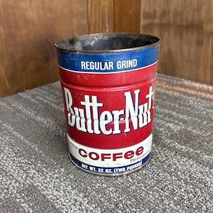以下が含まれることがあります： 赤、白、青のデザインが施された、ヴィンテージのバターナットコーヒー缶。「REGULAR GRIND」の文字が上部に、大きな白い文字で「Butter Nut」と書かれています。「The Coffee Delicious」と「NET WT. 907グラム」も記載されています。