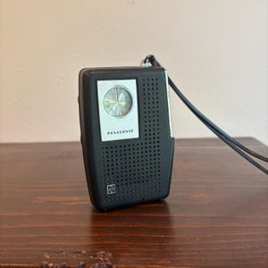 May include: A black Panasonic portable transistor radio with a perforated speaker grille, a silver-toned circular tuning dial, and a black wrist strap attached to the top. The device features a small square logo at the bottom left corner.