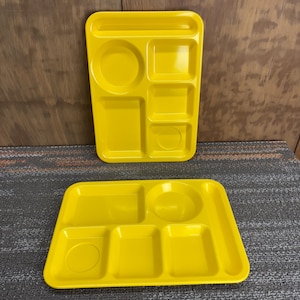 May include: Two bright yellow cafeteria trays. The trays have multiple compartments of various shapes and sizes, including a circular section for a drink and rectangular sections for food. The trays are made of a durable plastic material.