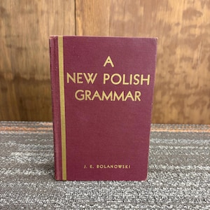 May include: A maroon-colored book titled "A New Polish Grammar" with gold lettering. The author's name, J. E. Bolanowski, is printed at the bottom. The book has a gold spine and is set against a wooden background.