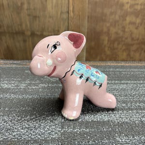 May include: A pink ceramic elephant figurine with a painted design on its back. The elephant has a smiling face, a red heart-shaped ear, and a blue and green design on its back. The figurine is on a gray surface.