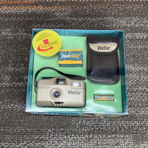 May include: A vintage Vivitar camera kit in a box. The kit includes a beige Vivitar camera, a black camera case, a roll of Vastcolor film, and two batteries. The box has a green interior and a yellow sticker that says "afterschool".