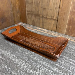 May include: A rectangular wooden tray with a dark brown finish. The tray has a carved design in the center and a handle on each end. The tray is sitting on a gray surface.