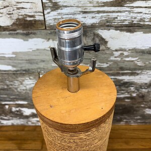 May include: A vintage table lamp with a cylindrical base and a wooden top. The base is wrapped in a textured, light brown material. The lamp features a silver-coloured socket and a black on/off switch. The lamp is designed for a standard light bulb.