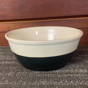 Puede incluir: Un cuenco de cerámica de dos tonos. La parte superior es blanquecina y la sección inferior es negra. El cuenco tiene una forma redondeada y un acabado liso y brillante. La sección negra tiene un aspecto texturizado y tejido. Adecuado para servir o exhibir.