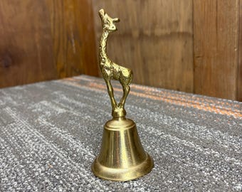 May include: A brass bell with a giraffe figurine on top. The giraffe is standing with its head raised. The bell has a flared base and a smooth, polished surface. The bell is a decorative item.