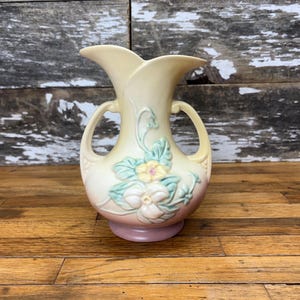 May include: A vintage ceramic vase with two handles, featuring a floral design with yellow and white flowers and green leaves. The vase has a light yellow and pink glaze.