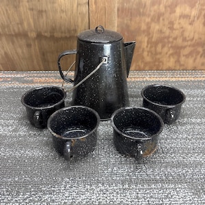 Peut inclure: Une cafetière en émail noir moucheté avec un couvercle, une poignée et quatre tasses assorties. L'ensemble est disposé sur une surface grise avec un fond en bois. La cafetière a un bec et une poignée en fil de fer.