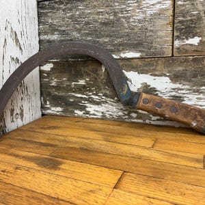 May include: A rusty metal and wooden hand sickle with a curved blade and a wooden handle. The sickle is resting on a wooden surface.