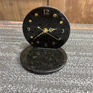 Puede incluir: Un reloj de escritorio de mármol negro con detalles dorados. La esfera del reloj es redonda con marcadores y manecillas doradas. La base también es de mármol negro, sobre una alfombra gris.