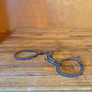 May include: A pair of antique iron shackles featuring two circular cuffs connected by a short, heavy-duty metal chain. The metal surface displays a dark, weathered, and rusted patina throughout, indicating age and extensive historical use.