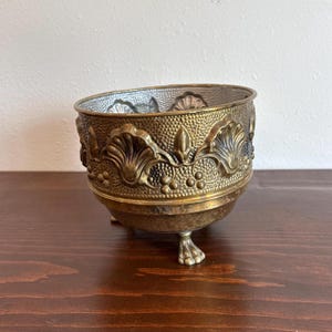 May include: A vintage brass bowl with ornate detailing. The bowl features a textured surface and is adorned with embossed floral and leaf patterns. It rests on three small, claw-shaped feet, adding to its antique aesthetic. The interior has a silver-colored finish.