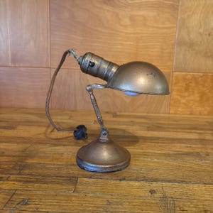 May include: A metallic desk lamp with a rounded shade, an adjustable articulated arm, and a circular base. The lamp features a weathered bronze-toned finish and includes a frayed fabric-covered electrical cord extending from the back.