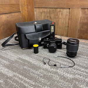 May include: A black Petri SLR camera with an attached flash unit, a separate JCPenney lens, a black leather carrying case with a shoulder strap, a small yellow-capped film canister, and a thin black shutter release cable with a metal tip.