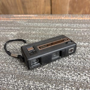 May include: A black Kodak Tele-Instamatic 608 camera with a wrist strap. The camera has a rectangular shape with a brown accent and the text "KODAK TELE-INSTAMATIC 608 MADE IN USA". The camera is on a gray surface.