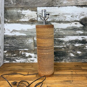 May include: A cylindrical table lamp base featuring a textured, tan-coloured corrugated cardboard exterior. The lamp includes a silver-toned metal socket fixture at the top and a black electrical cord extending from the base.