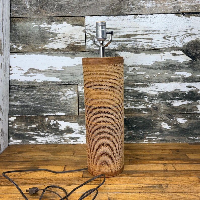 May include: A cylindrical table lamp base featuring a textured, tan-coloured corrugated cardboard exterior. The lamp includes a silver-toned metal socket fixture at the top and a black electrical cord extending from the base.
