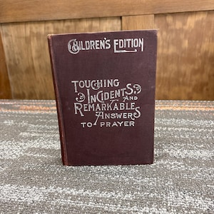 May include: A maroon-colored book titled "Children's Edition" with the text "Touching Incidents and Remarkable Answers to Prayer" in silver lettering. The book is standing upright on a patterned surface.