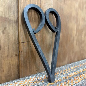May include: A handcrafted, dark grey metal heart-shaped wall decoration. The metal is forged with a textured finish, featuring two looped swirls at the top and a pointed base. The heart is leaning against a wooden surface.