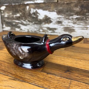 Puede incluir: Un recipiente negro de cerámica con forma de pato, pico dorado y collar rojo. El recipiente tiene asas pequeñas y un patrón decorativo. El pato está sobre una superficie de madera. Medidas en centímetros.