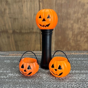 May include: Orange plastic Halloween decorations. A tall black stand with an orange pumpkin head, and two smaller orange pumpkin buckets with black handles and jack-o'-lantern faces. The decorations are on a gray and brown surface.