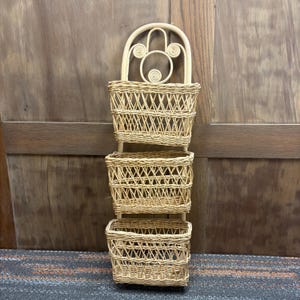 May include: A woven wicker wall organizer with three rectangular baskets. The top of the organizer has a decorative arch with spiral details. The baskets are attached vertically, offering storage and a natural aesthetic.