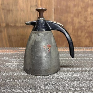 Puede incluir: Rociador de plantas de metal vintage con mango y boquilla negros. El rociador tiene un cuerpo cónico con un acabado gris descolorido y una pequeña pegatina colorida. La parte superior y el mango son negros.