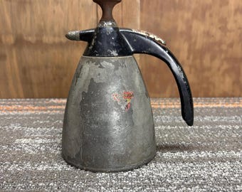 Puede incluir: Rociador de plantas de metal vintage con mango y boquilla negros. El rociador tiene un cuerpo cónico con un acabado gris descolorido y una pequeña pegatina colorida. La parte superior y el mango son negros.
