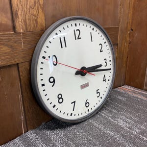 May include: A round, vintage wall clock with a white face and black numerals. The clock has a gray metal frame and a red second hand. The hands are black. The clock face has the brand name "Simplex".