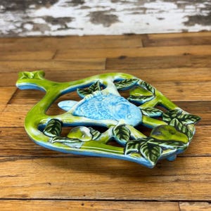 May include: A blue and green ceramic hot pad shaped like a tree branch with a bird and pears.